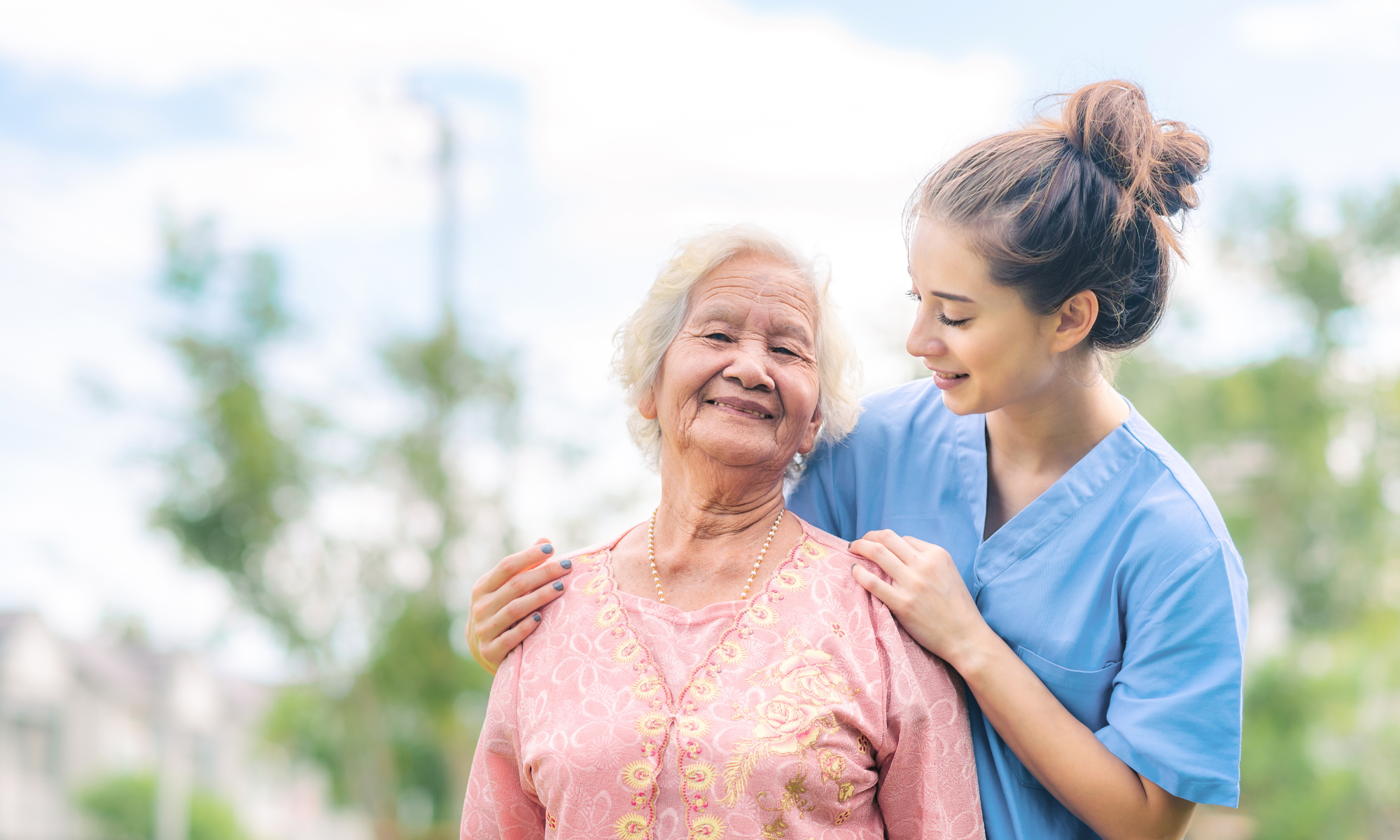 Caregiver and patient standing outdoors during daytime looking at eachother. Both are smiling.